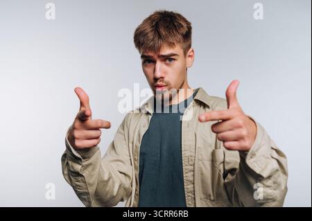 Ein junger Mann steht vor einem hellgrauen Hintergrund und schlägt eine verspielte Pose mit den Händen und macht Fingerpistolen. Er hat einen stilvollen Haarschnitt und ist zum Entspannen geeignet Stockfoto