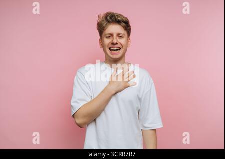 Junger Mann in einem weißen T-Shirt, lächelt mit Hand auf der Brust vor rosa Hintergrund. Stockfoto