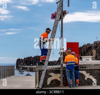 Stadtarbeiter in orangen Uniformen, die eine Straßenbeleuchtung an einer Strandpromenade in Camara de Lobos Madeira mit einer Leiter installieren Stockfoto