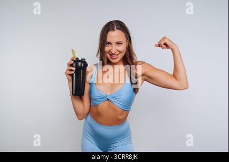 Frau in blauer Activewear, die Bizeps beugt und eine Shaker-Flasche hält. Stockfoto