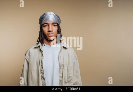 Junger Mann mit geflochtenen Haaren, der ein silbernes durag und beiges Hemd auf neutralem Hintergrund trägt. Stockfoto
