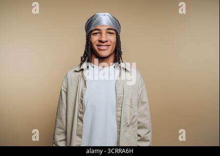 Junger Mann mit geflochtenen Haaren, der einen durag und legere Kleidung vor beigefarbenem Hintergrund trägt. Stockfoto