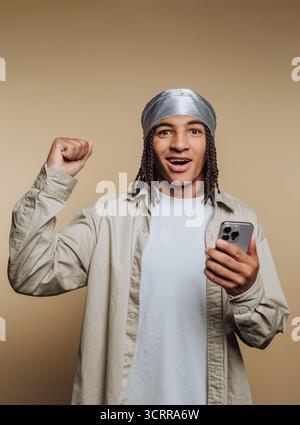 Aufgeregter junger Mann mit geflochtenen Haaren, der ein Smartphone hält, einen durag und lässige Kleidung trägt, vor einem beigen Hintergrund. Stockfoto