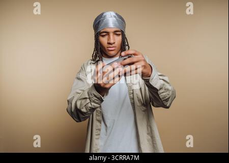 Junger Mann mit Zöpfen, der einen durag trägt, konzentriert sich auf ein Smartphone. Stockfoto