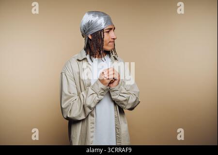 Junger Mann in lässiger Kleidung mit silbernem durag vor beigefarbenem Hintergrund. Stockfoto