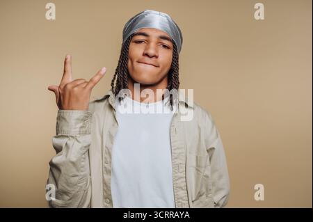 Junger Mann mit Zöpfen, der eine durag und legere Kleidung trägt, eine Handgeste vor einem beigen Hintergrund macht. Stockfoto