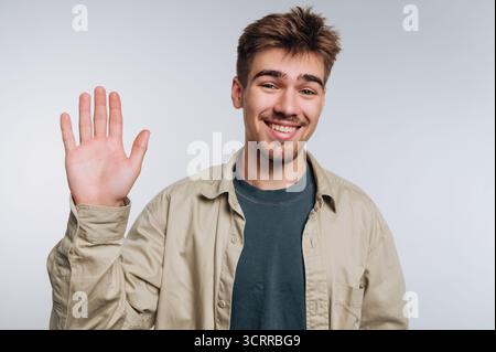 Junger Mann lächelt und winkt vor einem schlichten Hintergrund in die Kamera. Stockfoto