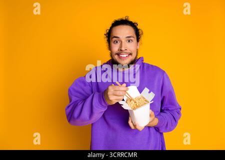 Junger Mann in einem violetten Hoodie, der Nudeln zum Mitnehmen mit Essstäbchen vor einem leuchtend gelben Hintergrund genießt Stockfoto