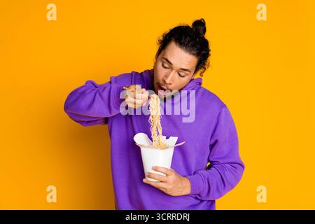 Porträt eines jungen Mannes in lässigem Hoodie, der Nudeln vor hellgelbem Hintergrund isst und Spaß und Lebensstil zum Ausdruck bringt Stockfoto