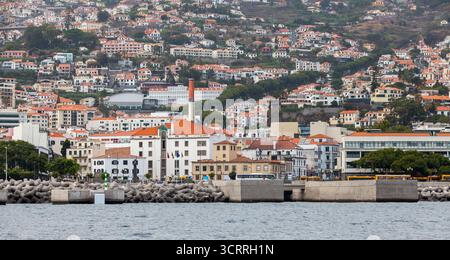 Blick aufs Meer auf Praca da Autonomia, Madeira, Portugal. Ein belebtes Uferviertel in einer Küstenstadt zeigt einen Hafen, einen Wellenbrecher und einen Hügel voller Berge Stockfoto