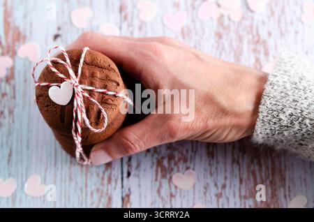 Eine weibliche Hand, die einen Stapel von gestapelten Schokoladenkeksen schnappt, gebunden mit einer roten und weißen Schnur und kleinen rosa Herzen auf hölzernem hellblauen Hintergrund. Stockfoto