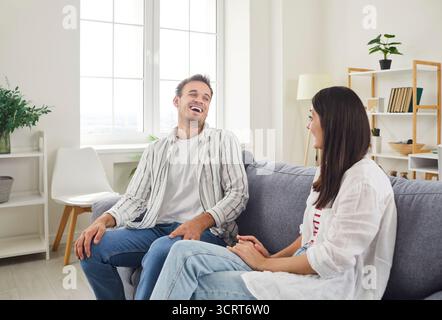 Glückliches junges Familienpaar, das auf einem gemütlichen Sofa sitzt, lacht und lustige Momente zu Hause genießt. Stockfoto
