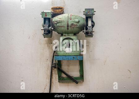 Eine grüne, an einem blauen Rahmen angebrachte industrielle Schleifmaschine steht an einer schmutzigen Wand in einer alten Werkstatt Stockfoto