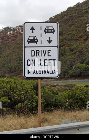 Straßenschild „Drive on Left in Australia“ zwischen Gras, Sträuchern und einem Berg mit Bäumen Stockfoto
