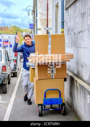 Junger Liefermann, der mehrere große Kisten auf einem Trolley mit Siegeszeichen beim Fotografieren zermürbt hat - Loches, Indre-et-Loire (37), Frankreich. Stockfoto