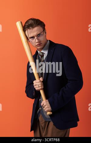 Ein trendiger Teenager steht selbstbewusst und hält einen Baseballschläger in einem leuchtenden orangefarbenen Raum. Stockfoto