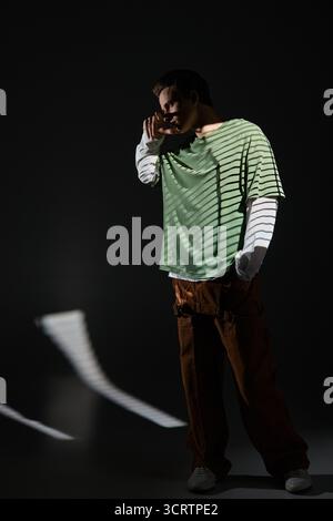 Ein junger Mann steht selbstbewusst in einem dunklen Raum und trägt trendige Kleidung, die die Jugendkultur widerspiegelt. Stockfoto