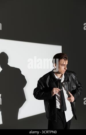 Ein junger Mann in einer Lederjacke steht selbstbewusst und zeigt die Jugendkultur in einem trendigen Studio-Licht. Stockfoto
