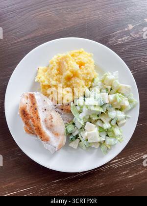 Nahrhafte Mahlzeit mit Hähnchenfilet, Maisbrei und frischem Salat wird auf dem Teller serviert. Das Geschirr steht auf einem rustikalen Holztisch Stockfoto