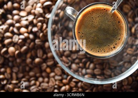 Detail einer Tasse Kaffee mit einem Metalllöffel auf einer Glasplatte auf einem Stapel gerösteter Kaffeebohnen. Draufsicht. Stockfoto