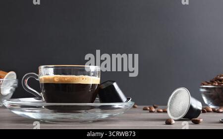 Espresso-Glas-Tasse auf einem Holztisch mit Aluminiumkapseln und Kaffeebohnen und einem isolierten schwarzen Hintergrund. Vorderansicht. Stockfoto