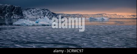 Eisberge schweben in einem teilweise gefrorenen Fjord bei Sonnenuntergang in der Nähe von Qeqertaq, Grönland. Bergkämme erstrahlen in sanftem Winterlicht über einer arktischen Meereslandschaft. Stockfoto