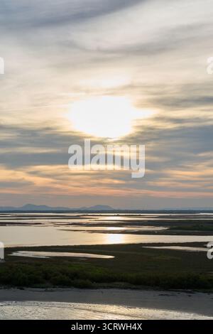 Die Sonne untergeht über einer ausgedehnten Feuchtlandschaft, erzeugt eine helle Reflexion auf dem ruhigen Wasser und zeigt eine ferne Bergsilhouette. Stockfoto