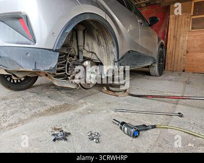 Tiefwinkelansicht eines silbernen Crossover-Fahrzeugs, das auf einem Wagenheber angehoben wurde und das Hinterrad entfernt ist, mit freiliegender Bremsbaugruppe und Werkzeugen in der Garage Stockfoto