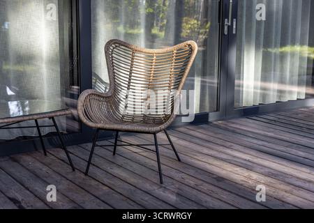 Das Bild zeigt einen wunderschön gestalteten Rattan-Stuhl auf einer Holzterrasse, begleitet von einem Glastisch, der moderne Wohnräume im Freien beleuchtet. Stockfoto