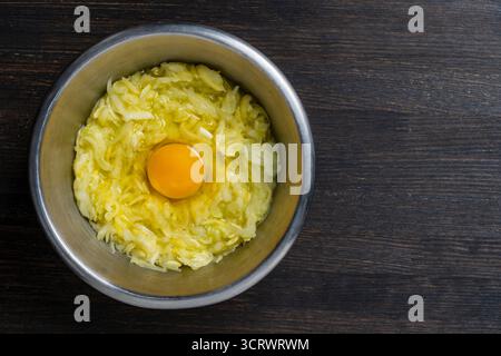 Geriebene Zucchini mit einem Ei in einer Metallschale auf einem Holztisch, Nahaufnahme, Draufsicht, Kopierraum. Rohstoffe für die Zubereitung von Gemüsewaffeln, Stockfoto