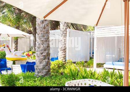 Luxuriöse Cabanas oder Zelt mit weißen Vorhängen und blauen Liegestühlen unter Palmen und Sonnenschirmen. Grüne Pflanzen und aufblasbares Einhornschweben sorgen für To Stockfoto