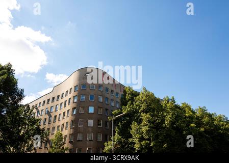 Das berühmte Bonjour Tristesse-Gebäude von Álvaro Siza in Berlin-Kreuzberg. Seine einzigartige geschwungene Fassade steht vor einem blauen Himmel, eingerahmt von grünen Bäumen. Stockfoto