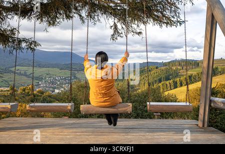 Eine Frau in einer gelben Jacke sitzt auf einer Schaukel mit Blick auf ein malerisches Bergtal mit Wäldern, Dörfern und bewölktem Himmel Stockfoto