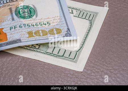 Die Ecke zweier sich überschneidender 100-Dollar-Scheine auf einer strukturierten braunen Lederoberfläche. Das Bild hebt Sicherheitsfunktionen und Druckdetails hervor. Stockfoto