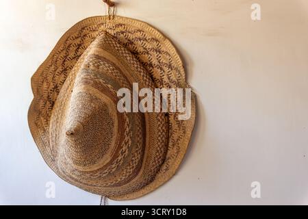 Traditioneller, handgewebter Sonnenhut mit Strohhalm und breiter Krempe, der an einem einfachen Wandhaken aufgehängt wird Stockfoto