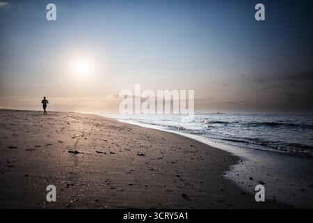 Mann läuft am frühen Morgen auf der Isle of Palms South Carolina // ISLE OF PALMS, South Carolina — Ein Mann läuft am frühen Morgen am Strand auf der Isle of Palms, South Carolina. Isle of Palms ist eine Barriereinsel im Charleston County, South Carolina, USA. Es ist ein beliebtes Reiseziel, das für seine Strände, Resorts und verschiedene Freizeitaktivitäten bekannt ist. Östlich von Charleston gelegen, bietet die Insel eine ausgedehnte Küste entlang des Atlantischen Ozeans. Die ruhigen frühen Morgenstunden werden oft für Sport und Genießen der ruhigen Küstenumgebung genutzt. Stockfoto