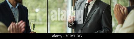 Kaukasischer Mann mittleren Alters im Anzug, der Champagner hält und unter einer Gruppe von Leuten steht, die während einer formellen Veranstaltung klatschen, teilweiser Blick auf verschiedene Erwachsene im Hintergrund Stockfoto