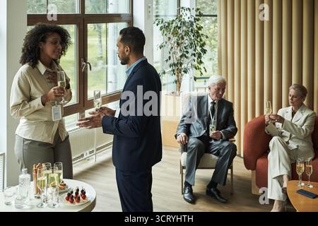 Schwarze Frau mittleren Alters und kaukasischer Mann mittleren Alters stehen und sprechen, während sie Champagner halten, kaukasischer Senior-Mann und kaukasische Senior-Frau sitzen und trinken im Hintergrund Stockfoto
