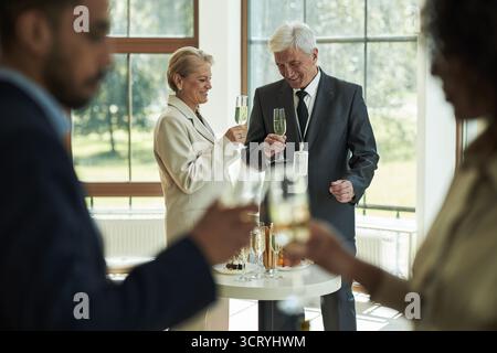 Kaukasische Frau mittleren Alters und kaukasischer Seniorenmann stehen zusammen, halten Champagner und lächeln während einer Geschäftsveranstaltung, während zwei verschwommene Menschen im Vordergrund stehen Stockfoto