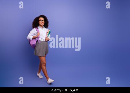Positive Frau mit Afro-strukturierten Haaren, die Notebooks halten und einen Rock mit einem lässigen Hemd vor einem lila Hintergrund tragen Stockfoto