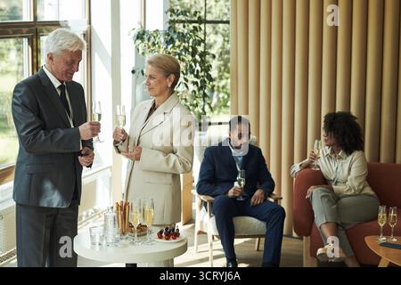 Senior-kaukasischer Mann und Senior-kaukasische Frau stehen und halten Champagner-Gläser, sprechen neben Tisch mit Getränken, junge Schwarze und junger Mann aus dem Nahen Osten sitzen und trinken Stockfoto