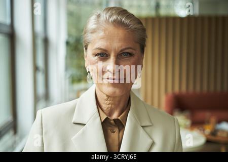 Porträt einer kaukasischen Frau mittleren Alters, die selbstbewusst lächelt, während sie in die Kamera blickt, drinnen mit kurzen blonden Haaren steht und Ohrringe trägt, Hintergrund leicht verschwommen Stockfoto