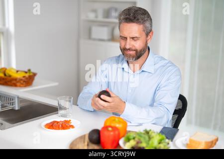 Der Mann isst morgens in der Küche. Mann mittleren Alters genießt frisches Essen am Küchentisch. Ein Mann isst Getreide und Obst in der Küche Stockfoto