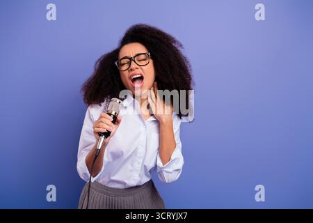 Junge Frau, die leidenschaftlich in ein Mikrofon singt, vor einem leuchtenden violetten Hintergrund in formeller Kleidung Stockfoto