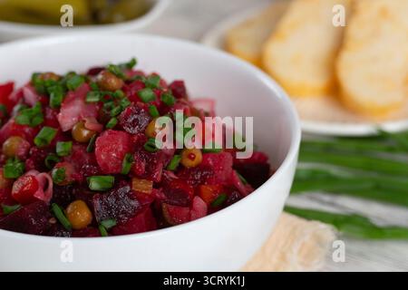 Hausgemachte russische Rote-Bete-Salat Vinaigrette, serviert in einer weißen Schüssel mit Brot, Gurken und grünen Zwiebeln. Traditionelles, gesundes vegetarisches Gericht Stockfoto