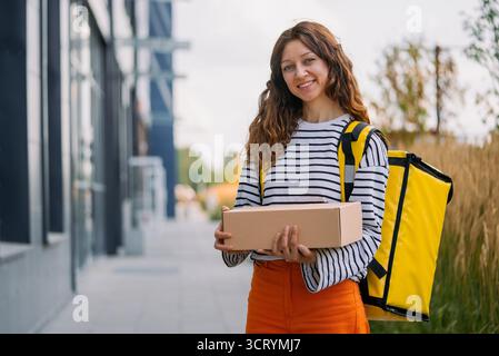 Lächelnde Zustellfrau mit einem gelben Thermorucksack und Paket in der Hand, die eine effektive Logistik auf der letzten Meile für schnellen Transport von Lebensmitteln und Paketen darstellt Stockfoto