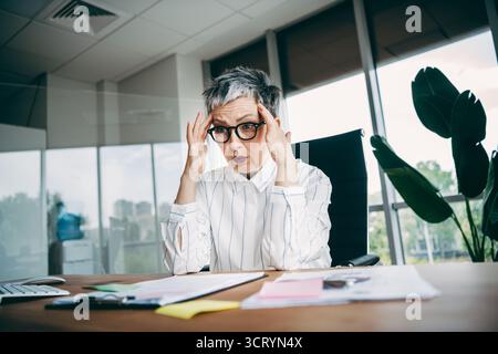 Ausgebildete Geschäftsfrau in stilvollem Büroumfeld, gestresst am Schreibtisch mit Papieren und Computer im professionellen Arbeitsbereich Stockfoto