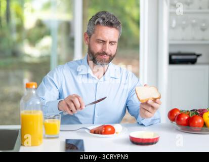 Der Mann isst morgens in der Küche. Der Mann aus den 40er Jahren genießt frisches Essen am Küchentisch. Ein Mann isst Getreide und Obst in der Küche. Guy genießt es Stockfoto