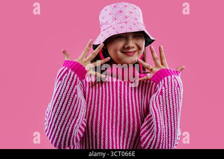 Verspielte asiatische Frau in einem rosa gemusterten Eimerhut und gestreiftem Grobpullover, lächelt und zeigt auf die Kamera vor einem leuchtend rosa Studio-Hintergrund. Stockfoto