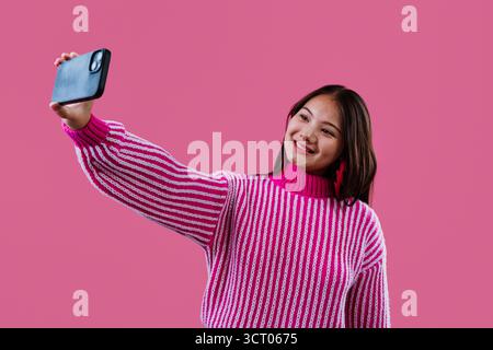 Eine junge Frau lächelt und macht ein Selfie mit ihrem Smartphone, gekleidet in einem gestreiften rosa-weißen Pullover, der moderne Kommunikation und soziale Medien reflektiert Stockfoto